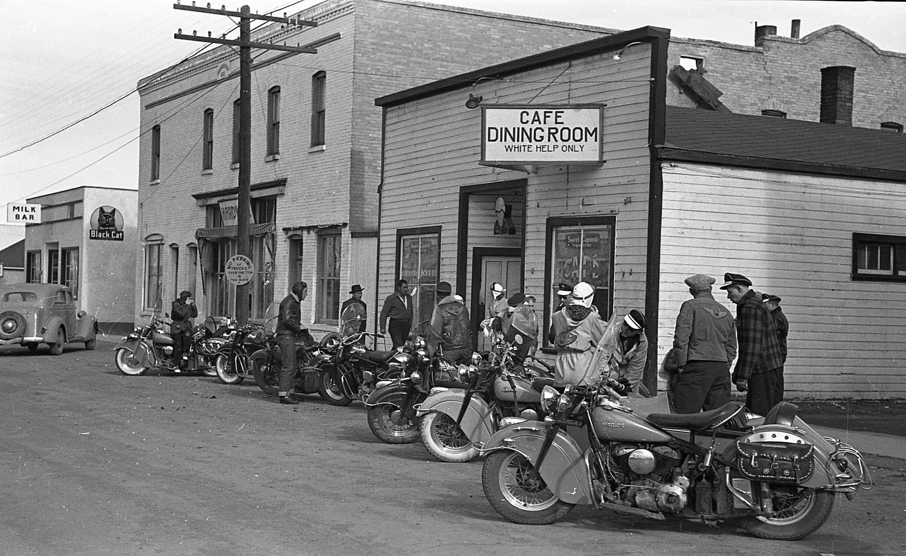 Ace-Hy motorcycles lined along a prairie main street outside a cafe whose sign reads Cafe Dining Room, White Help Only