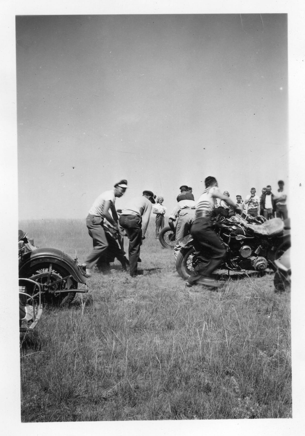 Ace-Hy riders scrambling to their motorcycles at a race start in a grassy field as a crowd looks on